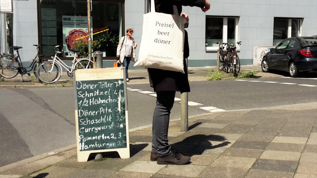 Frau auf Straße mit Jutebeutel, auf dem steht: Preiselbeerdöner, neben einem Aufsteller mit Döner-Angeboten